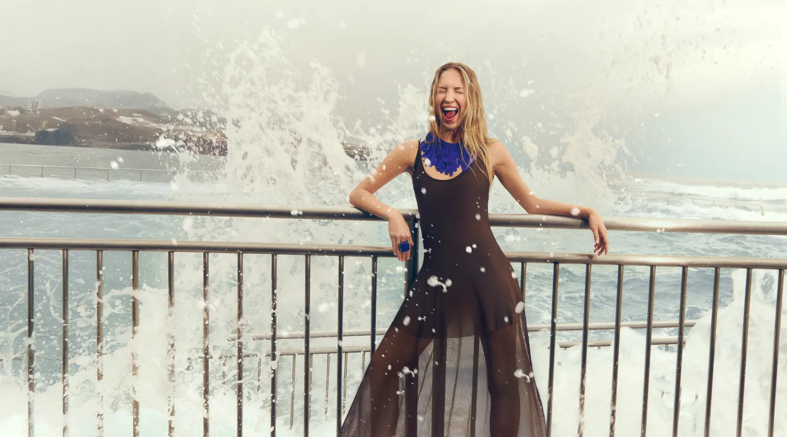 Vogue Spain // Lila Moss — A woman wearing a blue necklace is laughing while standing on a railing near a body of water.